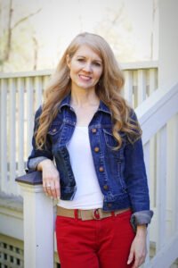 Shannon Bowie of Maple Cottage Designs in blue-jean jacket with white top Red Jeans and brown belt standing at foot of porch steps